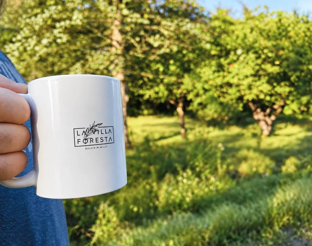 A hand holding a Villa Foresta branded mug with a green orchard in the background